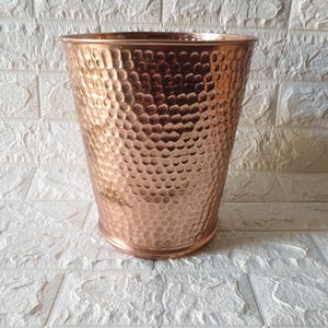 May include: A copper-colored metal wastebasket with a hammered texture. The wastebasket is cylindrical with a slightly flared top and a rounded base. The surface has a dimpled pattern, reflecting light. The background is a white brick wall.