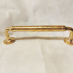 Könnte beinhalten: Ein goldfarbener Türgriff mit einem verzierten Design. Der Griff hat an jedem Ende eine abgerundete, flache Basis.