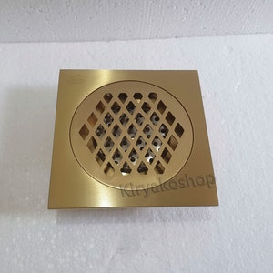 May include: A square, brass-colored floor drain with a circular, diamond-patterned grate. The drain has a brushed finish and is designed for water drainage in bathrooms or kitchens. The text "Kiryakosshop" is visible on the bottom right corner.