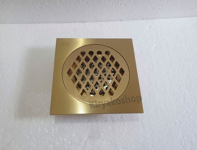 May include: A square, brass-colored floor drain with a circular, diamond-patterned grate. The drain has a brushed finish and is designed for water drainage in bathrooms or kitchens. The text "Kiryakosshop" is visible on the bottom right corner.