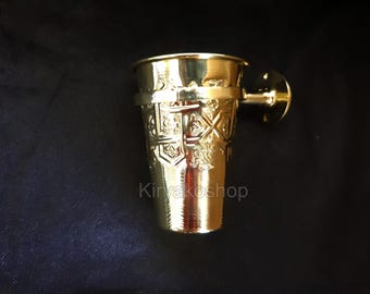 May include: Gold-toned metal cup with an ornate design and a wall mount. The cup has a round base and a cylindrical shape.