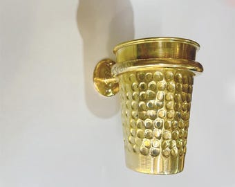 May include: A gold-colored metal toothbrush holder shaped like a thimble with a textured surface. The holder is mounted to a wall with a round base.