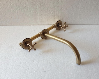 May include: A vintage-style brass faucet with a curved spout and two cross-shaped handles. The faucet has a warm, golden hue and is mounted on a horizontal bar. The design suggests a classic or antique aesthetic, suitable for bathroom or kitchen fixtures.