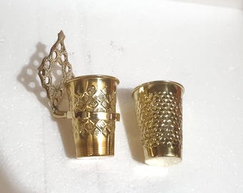 May include: Two gold-colored metal thimbles. One thimble is attached to a decorative, openwork metal wall-mount bracket with a diamond-shaped pattern. The other thimble features a textured, dimpled surface across its exterior body.
