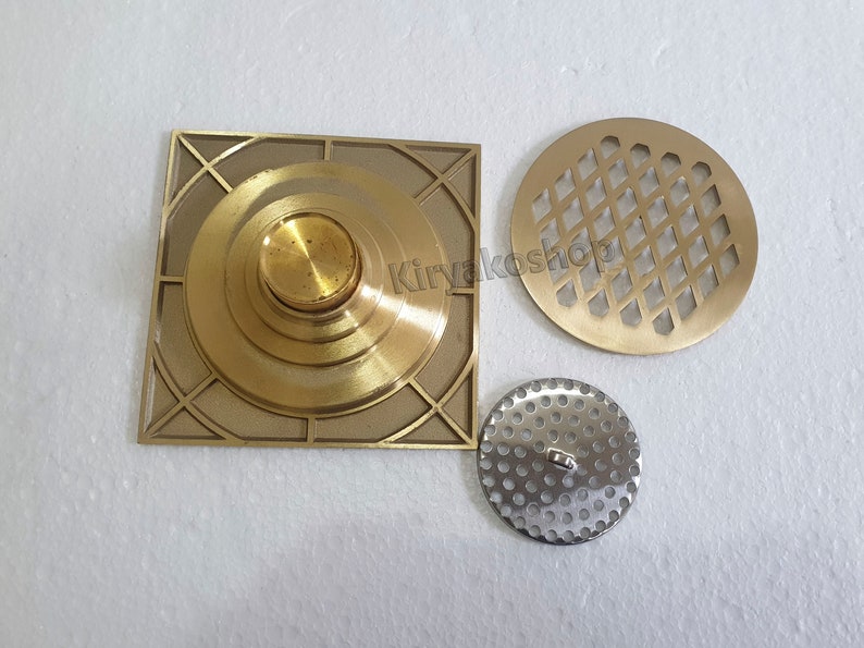 May include: A collection of brass and silver-toned metal drain covers. The square brass cover has a central knob and geometric design. A round brass cover features a diamond pattern, and a smaller, perforated silver-toned cover is also included.