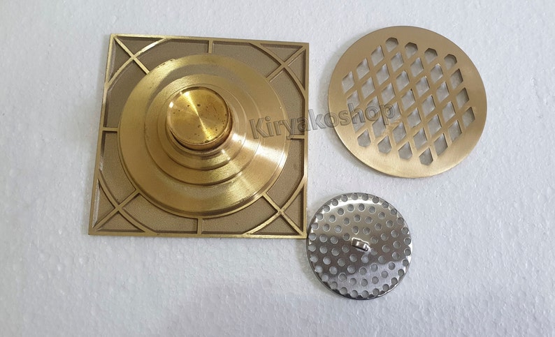 May include: A collection of brass drain covers and components. Includes a square drain cover with a central circular design, a round cover with a diamond pattern, and a smaller, perforated metal disc.