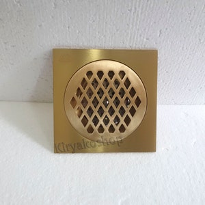May include: A square, brass-colored drain cover with a circular, diamond-patterned grate. The drain cover has a square frame and a central, diamond-shaped pattern for water drainage. The product is new.