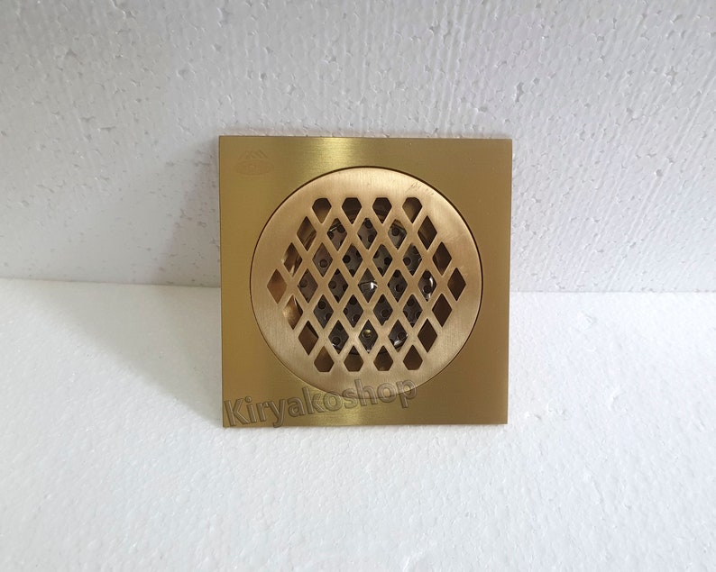 May include: A square, brass-colored drain cover with a circular, diamond-patterned grate. The drain cover has a square frame and a central, diamond-shaped pattern for water drainage. The product is new.