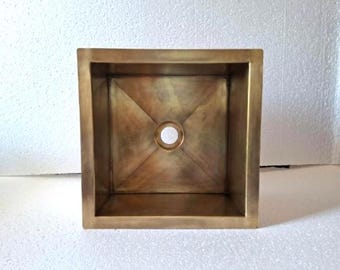 May include: A square-shaped sink with a metallic brass finish. The basin features a flat bottom with diagonal creases leading to a centered circular drain hole. The interior walls are vertical, and the rim is flat, creating a clean, geometric look.
