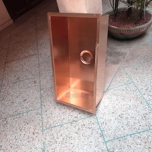Op de afbeelding: Een koperkleurige rechthoekige spoelbak met een cirkelvormig afvoergat. De spoelbak heeft een gepolijste afwerking en staat op een betegelde vloer. De merknaam "Kiryakosshop" is onderaan zichtbaar.
