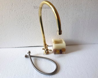 May include: A gold-colored faucet with a curved spout and cross-shaped handles. A braided metal hose and a small square candle with dried flowers are also present. The faucet is designed for a sink or basin.