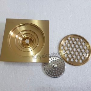 May include: A set of brass-colored drain components, including a square base with concentric circular grooves, a round perforated metal plate, and a decorative round grate with a diamond pattern. The components are designed for plumbing applications.