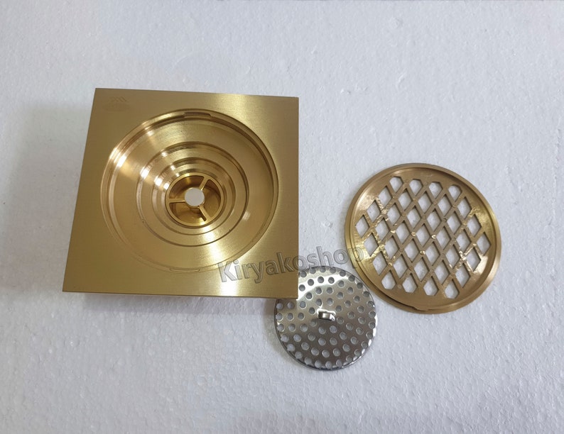 May include: A set of brass-colored drain components, including a square base with concentric circular grooves, a round perforated metal plate, and a decorative round grate with a diamond pattern. The components are designed for plumbing applications.