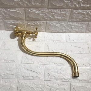 Può includere: Un rubinetto a muro curvo color oro con una maniglia a forma di croce.