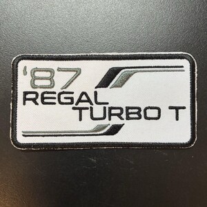 May include: A rectangular embroidered patch with a white background and a black border. The text reads '87 REGAL TURBO T' in black lettering. It features stylized black and grey graphic lines extending horizontally across the top and bottom.