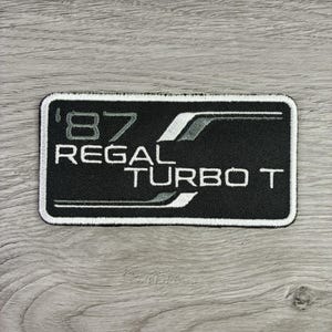 May include: A rectangular embroidered patch with a black background and a white border. The patch features the white text '87 REGAL TURBO T' with stylized white and grey graphic lines extending horizontally across the center of the design.