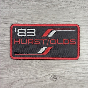 May include: A rectangular embroidered patch with a black background and a red border. The patch features the white text '83' and the red text 'HURST/OLDS' with stylized white and red graphic stripes flanking the text on the right and left sides.
