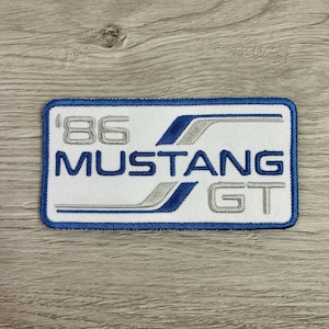 May include: Embroidered patch with a white background, blue border, and blue and silver accents. The patch features the text "'86 MUSTANG GT" in a stylized font, suitable for jackets or bags.