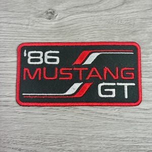 May include: Black rectangular patch with red border featuring the text "'86 MUSTANG GT" in white and red. The design includes red and white stripes. The patch is likely for a car enthusiast.