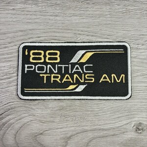 May include: A rectangular embroidered patch with a black background and a white border. The text '88 PONTIAC TRANS AM' is stitched in gold and white thread, accompanied by stylized gold and white horizontal lines across the design.
