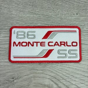 May include: A rectangular patch with a white background and a red border. The patch features the text "'86 MONTE CARLO SS" in silver and red. The design includes silver stripes and a red outline.