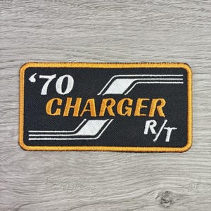 May include: A rectangular embroidered patch with a black background and a gold border. The text '70 CHARGER R/T' is stitched in white and gold thread, accompanied by white graphic lines that frame the text on this automotive-themed accessory.
