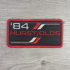 May include: A rectangular embroidered patch with a black background and a red border. The text '84' appears in white, followed by 'HURST/OLDS' in red. The design includes stylized white and red diagonal stripes across the black fabric surface.