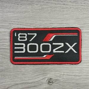 May include: A rectangular embroidered patch with a black background and a red border. The patch features the white text '87' above the text '300ZX' in a bold font. A stylized red and white graphic line runs horizontally across the design.