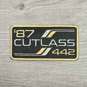 May include: A rectangular embroidered patch with a black background and a gold border. The text '87' and '442' appear in gold, while 'CUTLASS' is in white. Two stylized gold and white stripes run horizontally across the patch design.