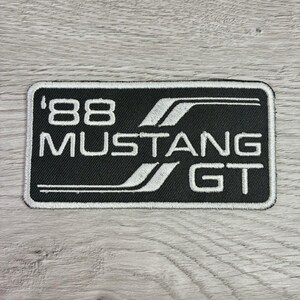 May include: Black rectangular patch with white embroidered text and design. The text reads "'88 MUSTANG GT". The design includes two curved white lines. The patch is likely for a car enthusiast.