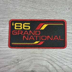 May include: Black rectangular patch with red border and text. The text reads "'86 GRAND NATIONAL" in yellow and red. The patch has yellow and red accent lines.