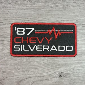 May include: A rectangular embroidered patch with a black background and a red border. The text '87' appears in white, followed by a red heartbeat line graphic. Below, the word 'CHEVY' is in red, and 'SILVERADO' is in white block lettering.