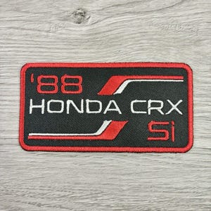May include: Black and red embroidered patch featuring the text "'88 HONDA CRX Si". The patch has a rectangular shape with rounded corners and white and red accent lines. The patch is on a gray wooden surface.