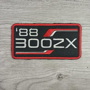 May include: A rectangular embroidered patch with a black background and a red border. The patch features the white text '88 300ZX' in a bold, stylized font, accented by two horizontal red stripes that frame the text at the top and bottom.