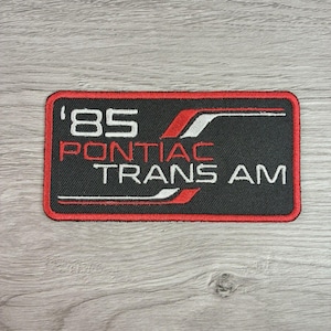 May include: A rectangular embroidered patch with a black background and a red border. The patch features the white text '85' and 'TRANS AM' with the red text 'PONTIAC' in the center. Red and white horizontal stripes accent the design.