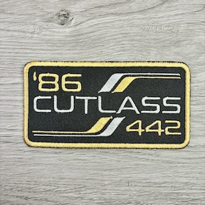 May include: Embroidered patch with a black background and gold trim. The patch features the text "'86 CUTLASS 442" in white and gold, with gold and silver accent stripes. The patch is rectangular with rounded corners.