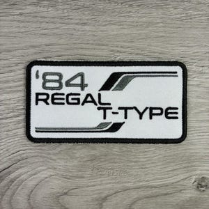 May include: A rectangular embroidered patch with a white background and a black border. The patch features the black text '84 REGAL T-TYPE' with stylized black and grey horizontal graphic lines extending from the letters across the white surface.