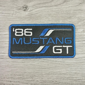 May include: Embroidered patch with a black background and blue border. The patch features the text "'86 MUSTANG GT" in white and blue. The design includes blue and white stripes.