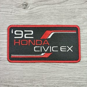May include: A rectangular embroidered patch with a black background and a red border. The text '92' and 'HONDA CIVIC EX' are stitched in white and red. Two horizontal red and white lines frame the text, creating a bold, graphic design.