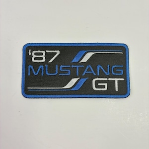 May include: A rectangular patch with a black background and a blue border. The patch features the text "'87 MUSTANG GT" in white and blue. The design includes stylized blue and white stripes.