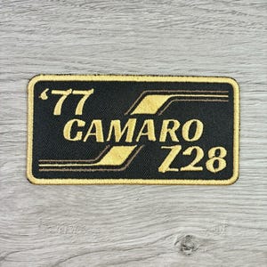 May include: Black rectangular patch with gold trim and lettering. The patch reads "'77 CAMARO Z28" in gold lettering. The patch has gold accent lines and is on a gray wood grain surface.
