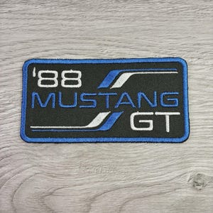 May include: A rectangular embroidered patch with a black background and a blue border. The text '88 MUSTANG GT' is stitched in white and blue. The design includes stylized blue and white horizontal lines flanking the text, creating a graphic emblem.