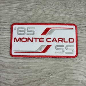 May include: A rectangular patch with a white background and red border. The patch features the text "'85 MONTE CARLO SS" in red and silver lettering. The design includes red and silver accent lines.