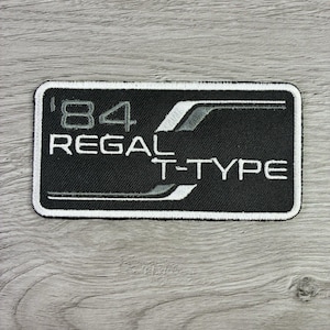 May include: A rectangular embroidered patch with a black background and a white border. The text '84 REGAL T-TYPE' is stitched in white, accompanied by a stylized white and grey graphic line that sweeps across the right side of the patch.
