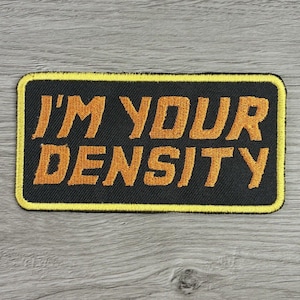 May include: A rectangular embroidered patch with a black background and a yellow border. The text "I'M YOUR DENSITY" is stitched in yellow, bold, sans-serif capital letters across two lines, centered within the patch's black field.