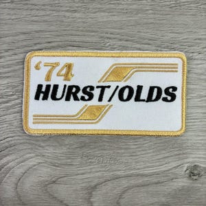 May include: A rectangular embroidered patch with a white background and a gold border. The patch features the gold text '74' and the black text 'HURST/OLDS' with gold decorative lines extending from the numbers and text across the white surface.