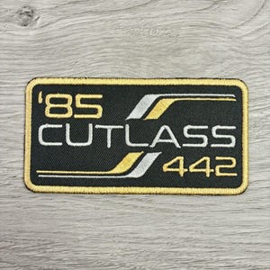 May include: A rectangular embroidered patch with a black background and a gold border. The text reads '85 CUTLASS 442' in white and gold lettering. The design includes stylized gold and white horizontal lines that accent the text.