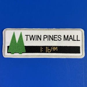 May include: A rectangular embroidered patch with a white background and a silver border. It features two green pine tree icons on the left, the black text TWIN PINES MALL, and a black horizontal stripe containing the gold text 1:16AM.