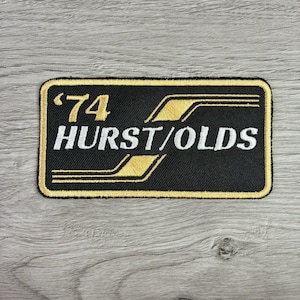 May include: A rectangular embroidered patch with a black background and a gold border. The patch features the gold text '74' and the white text 'HURST/OLDS' in a bold font. Two gold diagonal stripes frame the text, creating a racing-inspired design.