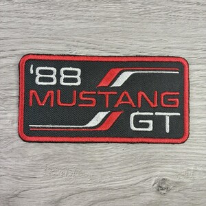 May include: A rectangular embroidered patch with a black background and a red border. The patch features the white text '88' and 'MUSTANG GT' with red and white graphic stripes. The design is oriented horizontally with clean, stitched edges.